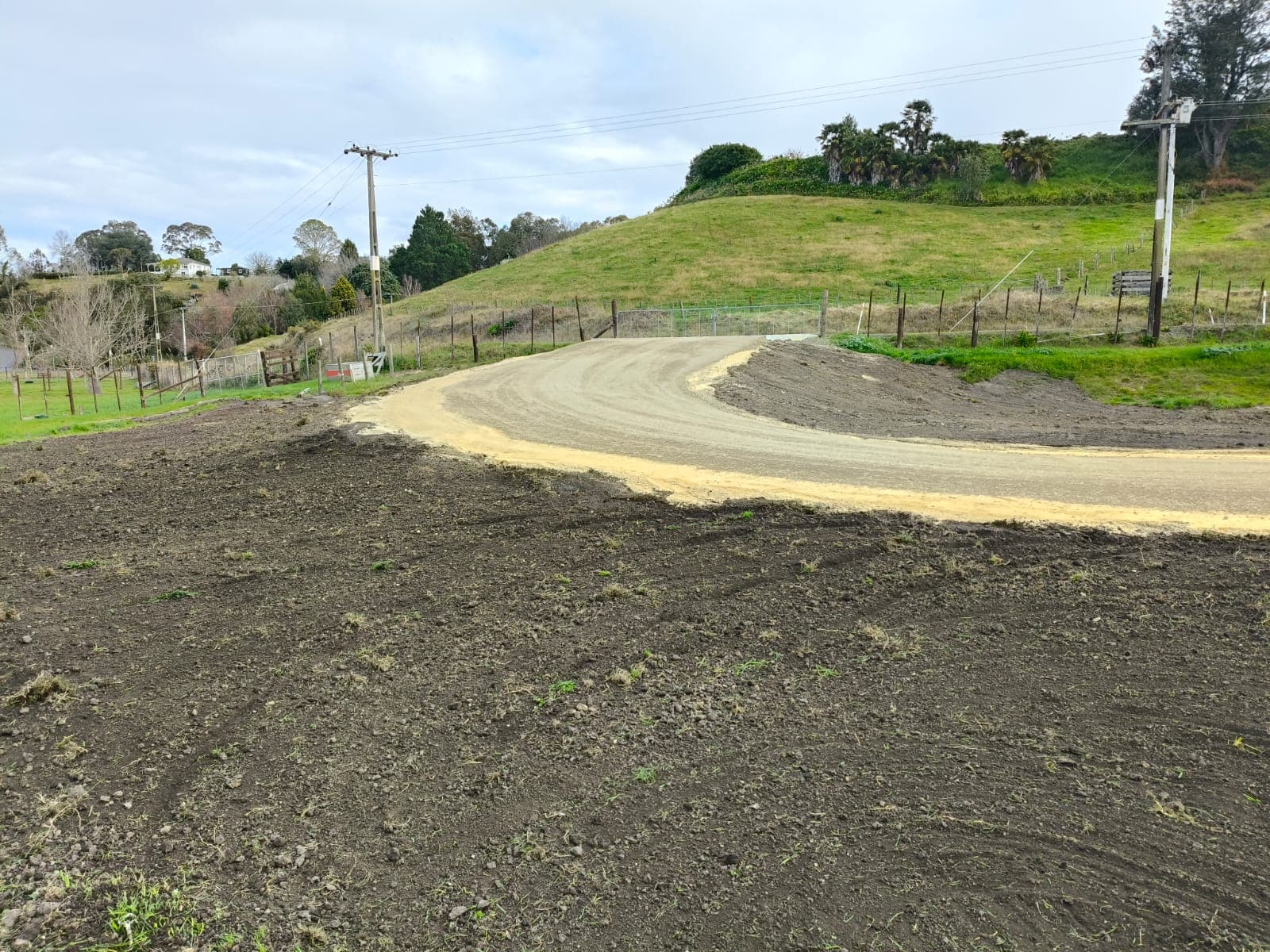Freshly graded curving gravel driveway through tilled earth with rolling green hills behind