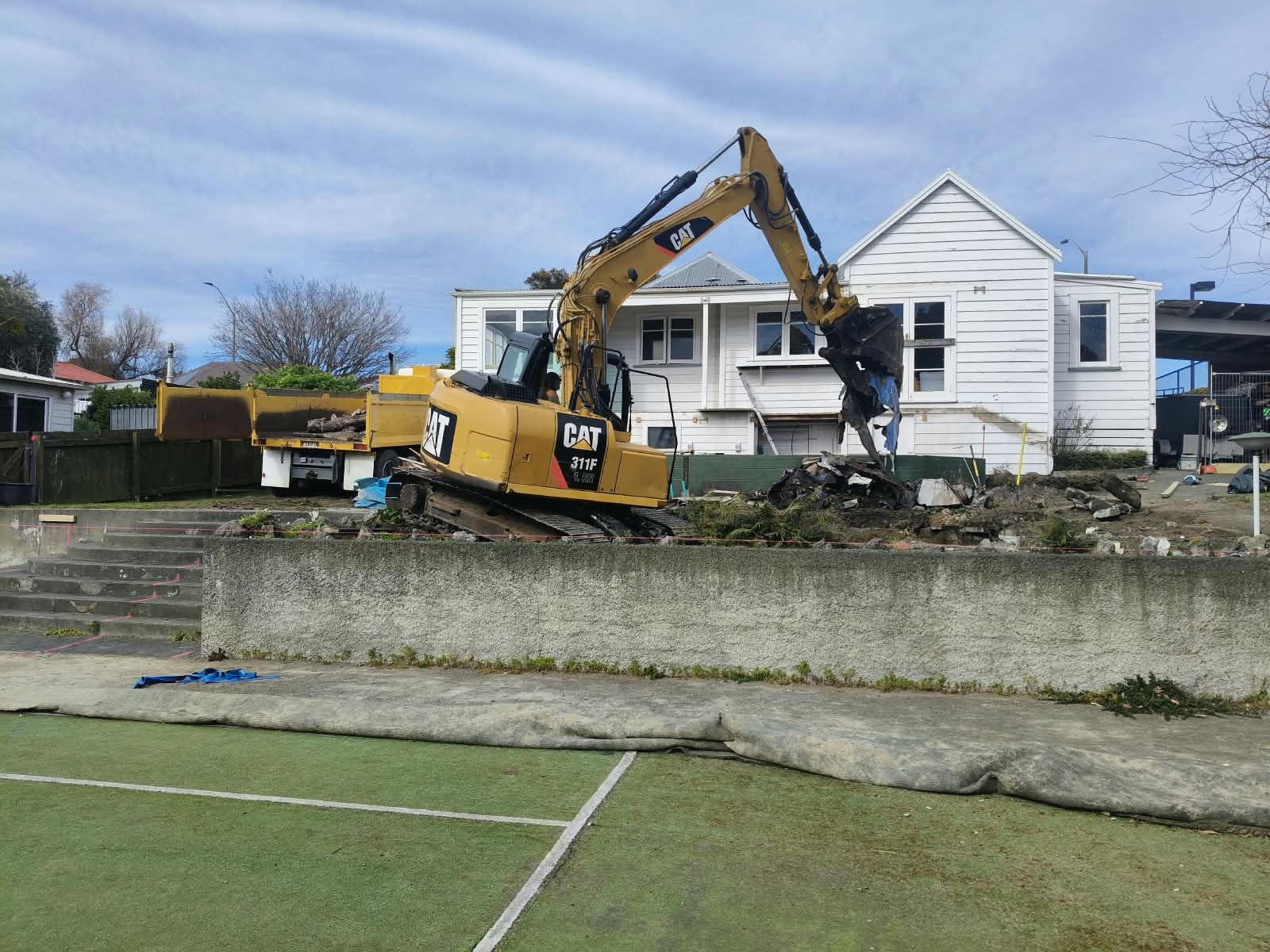 CAT excavator carrying out demolition and loading debris into a tip truck beside a house
