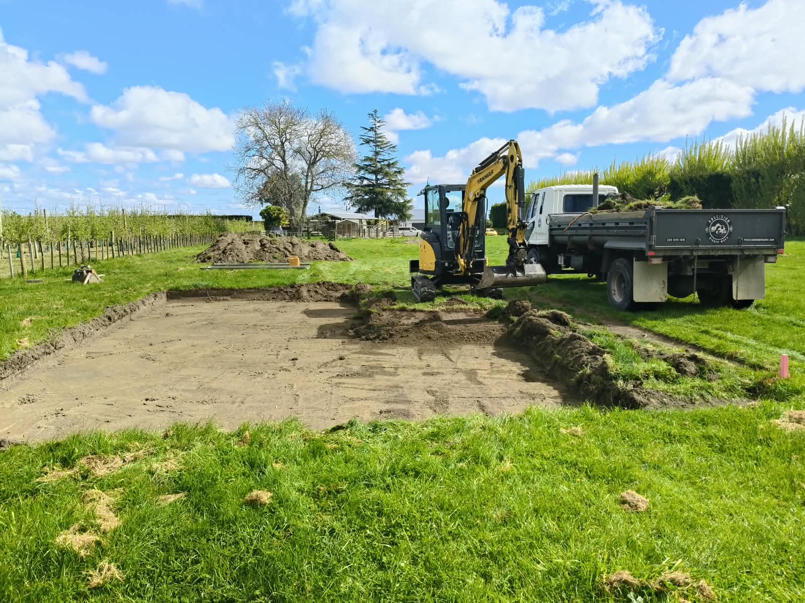 Wide foundation excavation beside an orchard with an excavator and truck
